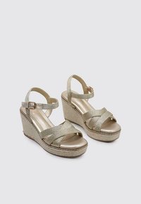 Le sandali wedge dorate presentano cinturini metallici incrociati e una suola intermedia in juta texturizzata. La soletta liscia e lucida aggiunge comfort e contrasto visivo.