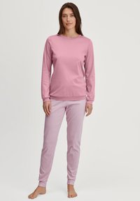 Vrouw die blootsvoets staat in een langarmige roze top en lichtroze gestreepte broek tegen een eenvoudige witte achtergrond.