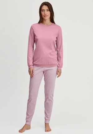Woman standing barefoot wearing a long-sleeve pink top and light pink striped pants against a plain white background.