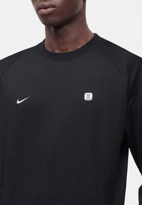 Νέος άντρας που φοράει μαύρο φούτερ Nike με άσπλο λογότυπο Nike και μικρό ορθογώνιο ταμπελάκι στο στήθος.