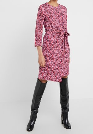 Kobieta w czerwono-różowej sukience o wzorze, sięgającej kolan, z wiązanym w pasie detalem i czarnych, wysokich na kolano butach na obcasie, stojąca na jednolitym tle.