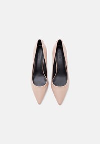 Ζευγάρι μπεζ μυτερές ψηλοτάκουνες γόβες με μαύρα πέλματα που φέρουν ανάγλυφο το λογότυπο "HUGO", εμφανιζόμενες από πάνω σε λευκό φόντο.