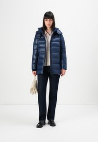 Marineblå pufferjakke med avtakbar hette, glidelås foran og horisontale sømmer. Modellen har på seg mørke jeans og holder en kremfarget håndveske.