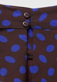 Brunt stof med blå ovale polka prikker, der har to mørke knapper ved taljen og en lille fold detalje under knapperne.