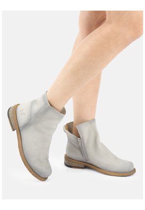 Persoon met lichtgrijze enkellaarzen met ritssluitingen aan de zijkant en houten zolen, staand op een witte achtergrond.