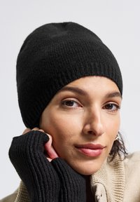 Beanie nera in maglia con texture a coste, caratterizzata da una vestibilità aderente. Il modello indossa guanti senza dita abbinati, che mostrano un materiale e un design simili.