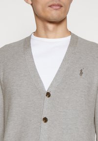 Ljusgrå cardigan med V-halsdesign, utrustad med två bruna knappar och en liten logotyp på vänster sida, över en vit T-shirt.