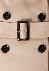 Tessuto del trench beige con una texture liscia, caratterizzato da grandi bottoni neri e una fibbia rettangolare in tartaruga sulla cintura.