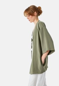 Olijfgroene, losse kimono-jas gemaakt van zachte stof, met brede mouwen en zijzakken, gedragen met een witte outfit eronder.
