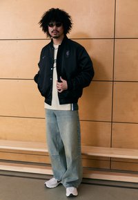 Musta satiini bomber-takki, jossa violetti logo, beige t-paita, leveät vaaleansiniset farkut ja valkoiset tennarit. Taustalla puupaneelit.