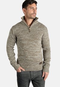 Maglione a maglia marrone con colletto alto e dettagli con zip, caratterizzato da un motivo testurizzato e polsini a costine, abbinato a jeans scuri.