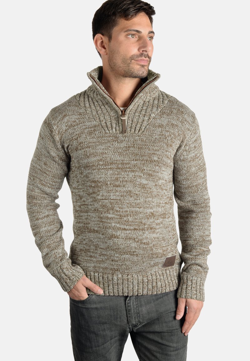 Maglione a maglia marrone con colletto alto e dettagli con zip, caratterizzato da un motivo testurizzato e polsini a costine, abbinato a jeans scuri.