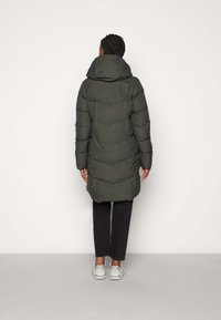 Zelený prošívaný kabát s kapucí, sahající ke kolenům, s prošívanými detaily a logovou nášivkou ve spodní části. Kombinován s tmavými kalhotami a světlými botami.