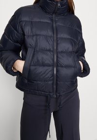 Námořnicky modrá prošívaná bunda s vysokým límcem, zapínáním na zip vpředu, viditelným horizontálním prošitím a elastickým lemem. Má boční kapsy a stahovací šňůrky na spodní části.