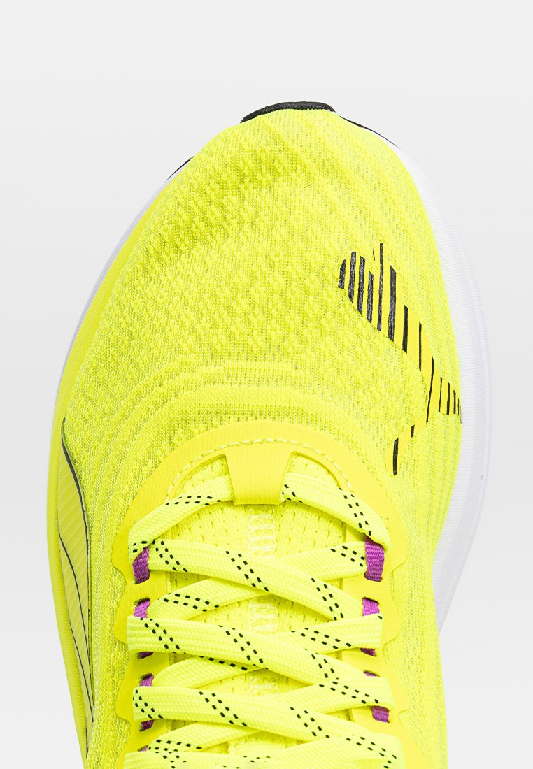 Jasnożółty sportowy but z teksturowaną cholewką z dzianiny, czarnymi paskami oraz fioletowymi detalami sznurowania. Posiada zaokrągloną białą podeszwę.