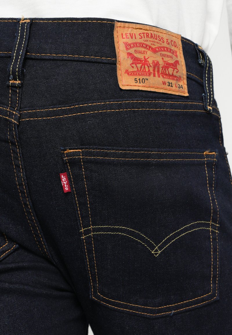 Jean en denim bleu foncé avec des coutures jaunes. Dispose d'une étiquette en cuir au dos portant l'inscription "Levi Strauss & Co." avec le numéro de modèle et le logo de la marque.