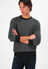 Grauer Sweater mit Fischgrätenmuster, rundem schwarzen Ausschnitt und Bündchen, ausgestattet mit einem kleinen Markenlogo auf der Brust. Kombiniert mit dunklen Jeans.