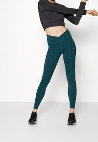 Teal legging tinggi dengan bahan yang halus dan elastis. Atasan lengan panjang hitam dan sepatu olahraga hitam melengkapi pakaian tersebut.