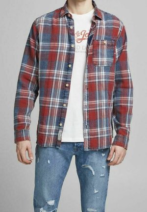 Man draagt een rood-blauw geruite blouse over een wit T-shirt met opdruk en verweerde blauwe spijkerbroek.