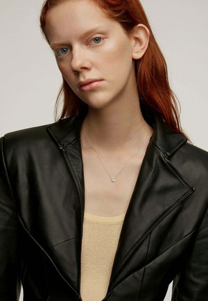 Blazer de cuero negro con diseño estructurado, superpuesto a una blusa de punto beige. Collar simple de plata con un colgante circular.