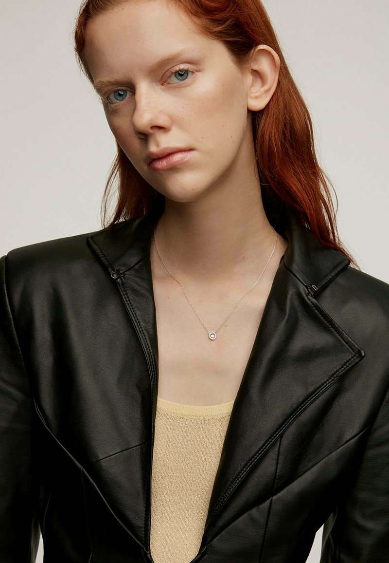Blazer de cuero negro con diseño estructurado, superpuesto a una blusa de punto beige. Collar simple de plata con un colgante circular.