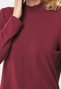 Burgunderfarbenes Langarmoberteil mit hohem Kragen, aus glattem Stoff hergestellt. Verfügt über eine figurbetonte Silhouette und subtile Nähedetails.