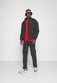 Sort bomberjakke med elastiske manchetter over en rød sweatshirt, parret med sorte bukser og sølvfarvede sneakers. En brun og sort bucket hat fuldender looket.