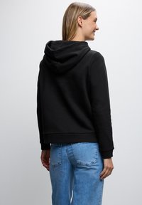 Felpa nera con una texture morbida, dotata di cappuccio con cordino e polsini a coste, abbinata a jeans azzurri chiari, leggermente usurati.