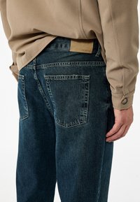 Tummansiniset denimfarkut klassisella leikkauksella, joissa on kaksi takataskua ja vaaleanruskea nahkapaikka vyötäröllä. Kangas on sileä.