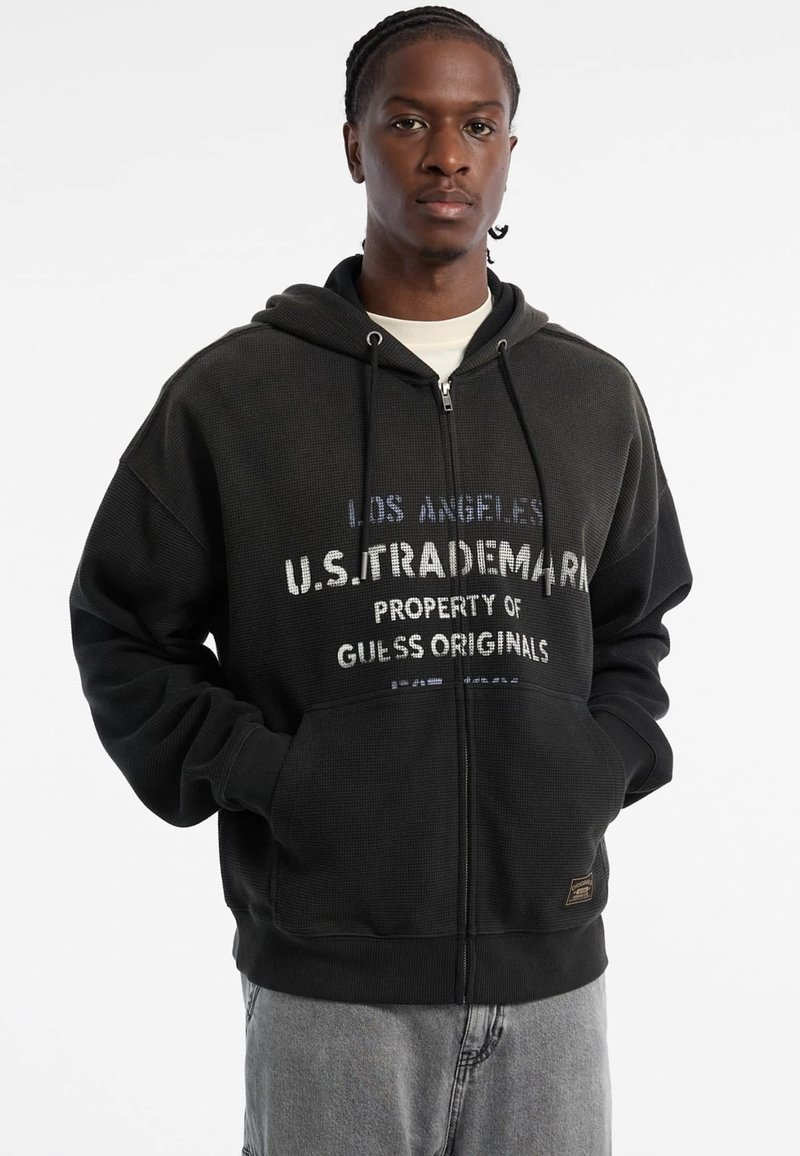Schwarze Zip-Hoodie mit strukturiertem Stoff, versehen mit weißem und blauem Logo-Text. Er hat eine Fronttasche und eine verstellbare Kapuze mit Zugband.