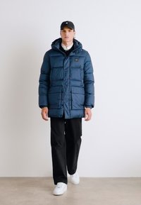Giacca a vento blu navy con cappuccio, caratterizzata da una zip frontale, cuciture orizzontali e due tasche laterali, abbinata a pantaloni neri e sneaker bianche.