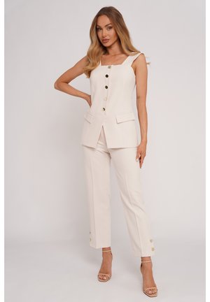 Kobieta o długich włosach, ubrana w kremowy, bezrękawnik z guzikami oraz dopasowane kremowe spodnie proste z guzikami przy kostkach, pozująca z jedną ręką na biodrze.