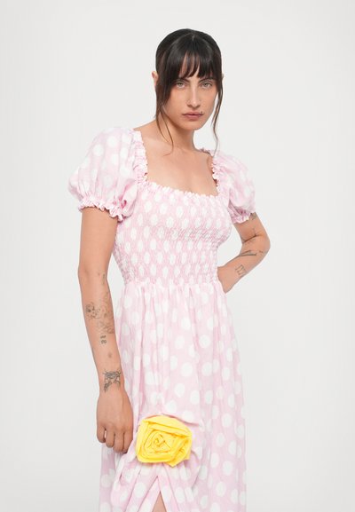 Lys pink kjole med hvide polkaprikker, puffærmer og smocked liv. Har en gul stofrose på裙et.