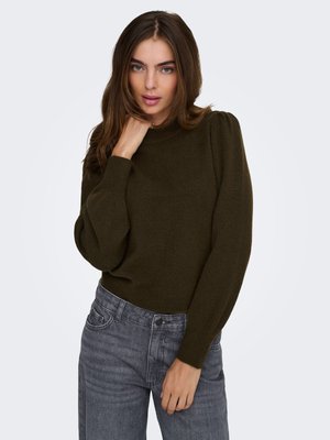 Jeune femme aux longs cheveux bruns portant un pull à manches bouffantes vert olive et un jean gris, posant avec une main près du cou.