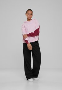 Roze en kastanjebruine hoodie met lange mouwen, vooraan een logo en een relaxte pasvorm, gecombineerd met losse zwarte broek en witte sneakers.