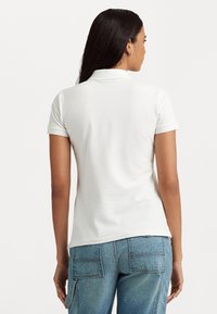 Λευκό κοντομάνικο μπλουζάκι πόλο με κλασικό γιακά, λείο υλικό και εφαρμοστή γραμμή, φορεμένο με ανοιχτό μπλε τζιν με τσέπες αγκώνα.