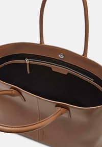 Brązowa skórzana torba typu tote z dwoma uchwytami, otwartym wierzchem i czarnym wnętrzem. Posiada zamykaną kieszeń i gładką teksturę. Minimalistyczny design.