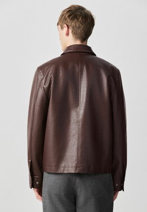 Junger Mann, der eine dunkelbraune Lederjacke mit langen Ärmeln und graue Hosen trägt, von hinten vor einem schlichten hellen Hintergrund.