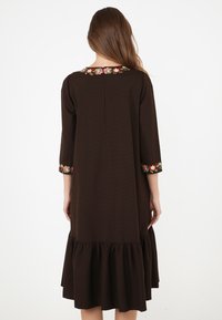 Kvinna med långt hår som bär en brun långärmad klänning med blommönsterbroderi på kragen och ärmsluten, står vänd bort med kjolens volang.