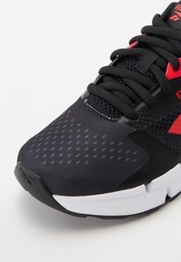 Črne atletske čevlje iz mrežaste in sintetične površine, s rdečimi detajli, teksturirano površino in belim gumijastim podplatom.