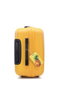 Valigia rigida gialla con superficie texturizzata, manico e zip neri. L'etichetta attaccata presenta un design geometrico di ananas.