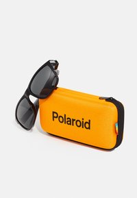 Fekete napszemüveg szürke lencsékkel, mellette egy fényes narancssárga téglalap alakú tok, amelyen a "Polaroid" vastag fekete betűkkel olvasható. A tok cipzárral záródik.
