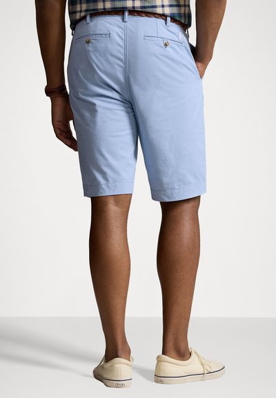 Ανοιχτό μπλε βαμβακερές κοντές παντελόνες με επίπεδο μπροστινό μέρος, δύο πίσω τσέπες με κουμπιά και λεία υφή. Φορεμένες με μπεζ χαμηλά παπούτσια.