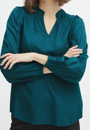 Vrouw draagt een tealkleurige blouse met lange mouwen, elastische manchetten, V-hals en subtiel patroon, armen gekruist en toont een gouden ring aan de linkerhand.