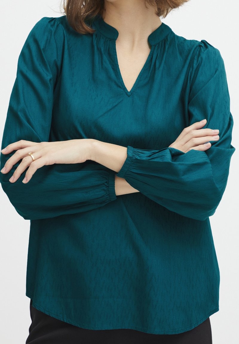 Femme portant un chemisier bleu sarcelle à manches longues avec des poignets élastiques, un col en V et un motif subtil, bras croisés montrant une bague en or à la main gauche.
