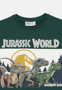 Σκούρο πράσινο κοντομάνικο μπλουζάκι που διαθέτει γραφικό από το Jurassic World με τρεις δεινόσαυρους και φόντο έναν κίτρινο κυκλικό ήλιο.