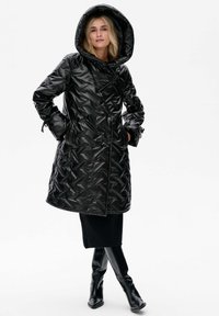 Zwarte gewatteerde jas met een driehoekig textuurpatroon, oversized capuchon, strikmanchetten en voorzakken, gedragen over een zwarte rok en kniehoge laarzen.