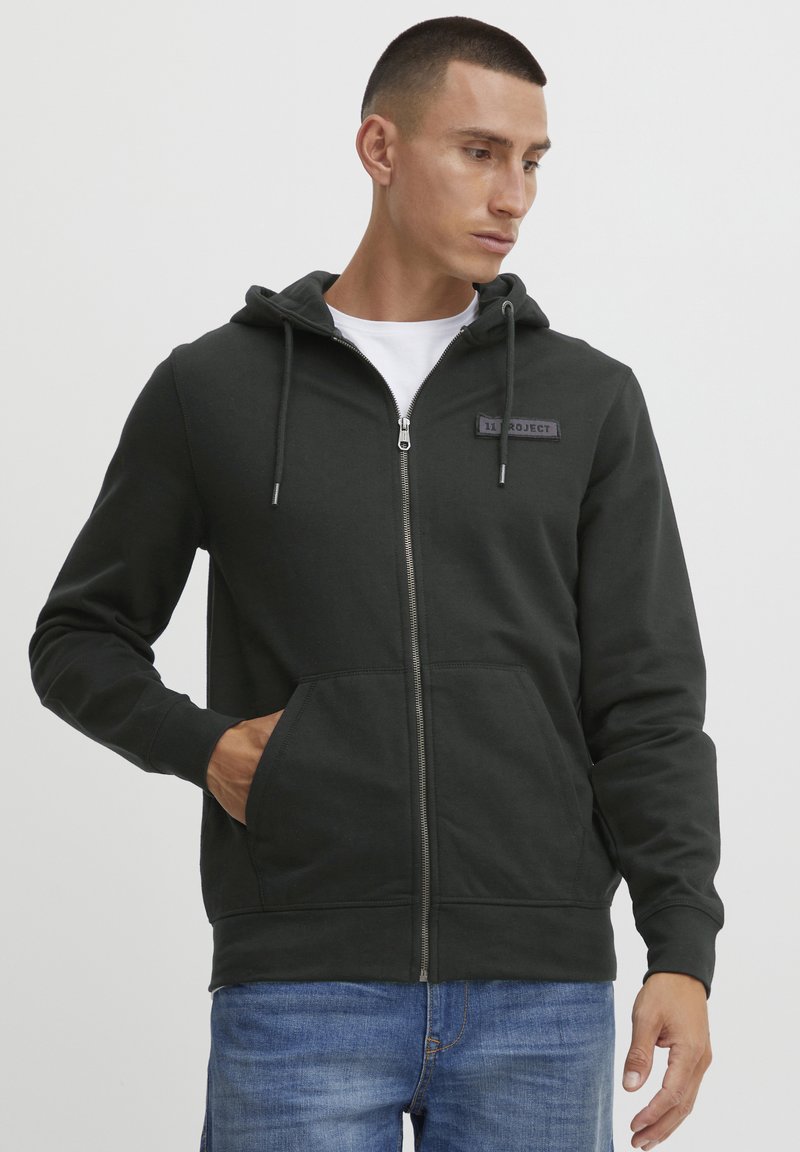 Sudadera con cremallera de color verde oscuro, con capucha ajustable, bolsillos frontales y una tela con textura. Presenta una etiqueta con "11 PROJECT" en el pecho.