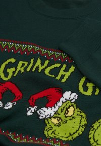 Pull vert en tricot avec l'inscription « GRINCH » en jaune, accompagné d'accents rouges et blancs. Le design inclut des personnages festifs portant des chapeaux de Père Noël.