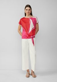 Rode en roze geblokte blouse met korte mouwen, geknoopt in de taille, gecombineerd met witte wijde broek en lichte sandalen.